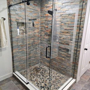Glass-enclosed shower with stone tile walls and pebble floor, black fixtures, and towel hanging nearby.