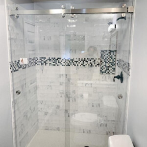 Glass-enclosed shower with sliding doors, white and gray tile walls, and a black-and-white mosaic accent strip.