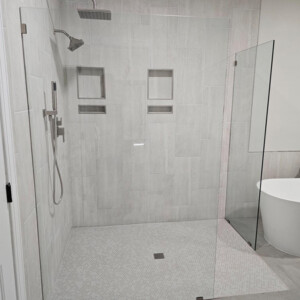 Modern walk-in shower with glass walls, rainfall showerhead, built-in shelves, and light gray tile surfaces.