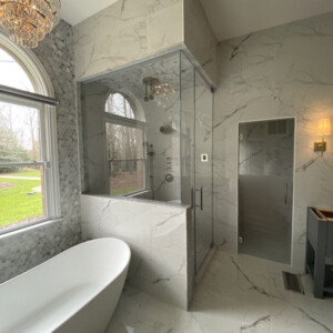 Modern bathroom with marble walls and floors, a glass shower, freestanding tub, and large window overlooking a lawn.